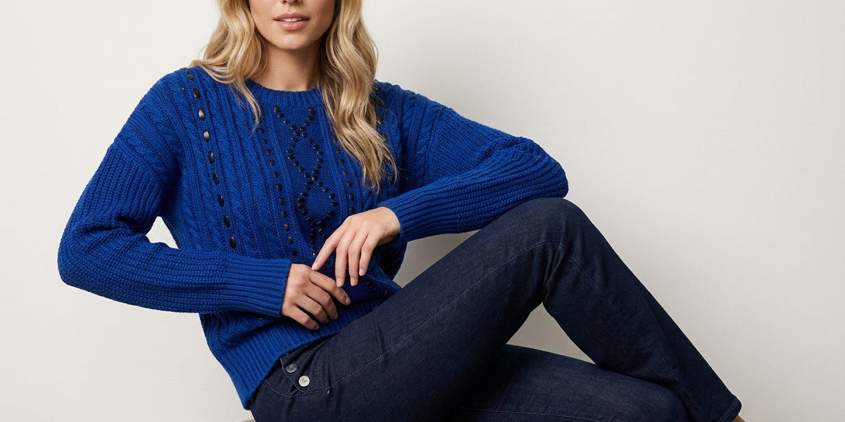 A woman sits on a round velvet stool wearing a textured royal blue cable-knit sweater and dark blue jeans against a plain light gray background.