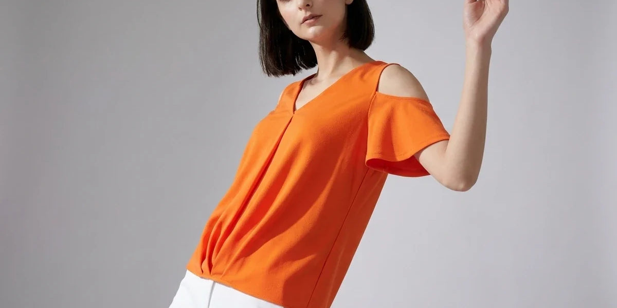 A woman modeling a bright orange cold-shoulder top with white pants, posed against a neutral gray studio background.