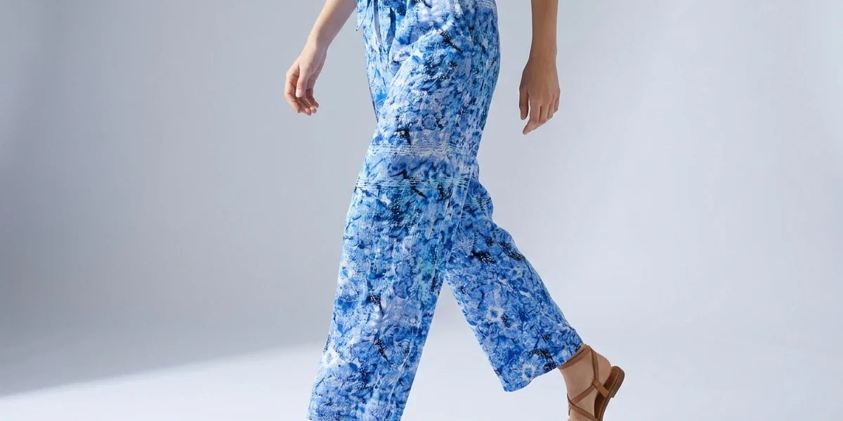 A model wearing blue and white abstract patterned wide-leg pants with a drawstring waist, styled with a cream top and brown leather sandals against a light gray studio background.