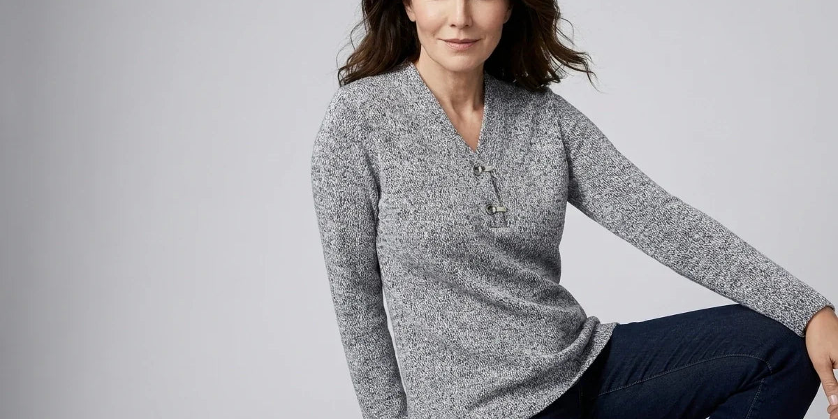 A woman wearing a gray marled long-sleeve henley sweater and dark blue jeans, posing against a light gray studio background.