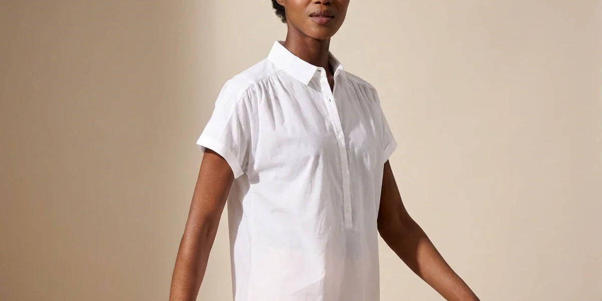 Black woman modeling a white short-sleeve collared button-up blouse with shoulder gathers and light pink pants in a studio setting.