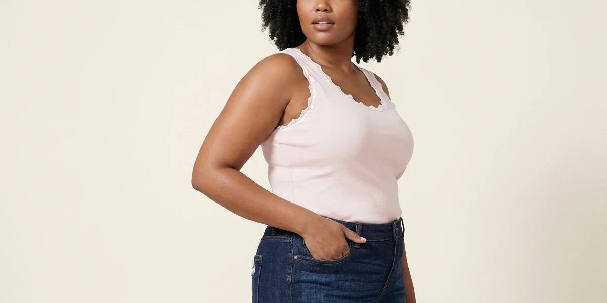 Curvy woman with curly black hair modeling a light pink lace-trimmed scoop-neck tank top tucked into dark wash jeans.