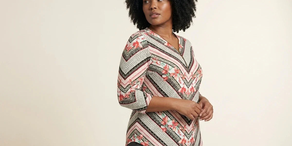 Curvy woman with curly black hair modeling a plus-size chevron and floral print three-quarter sleeve tunic top paired with grey trousers.