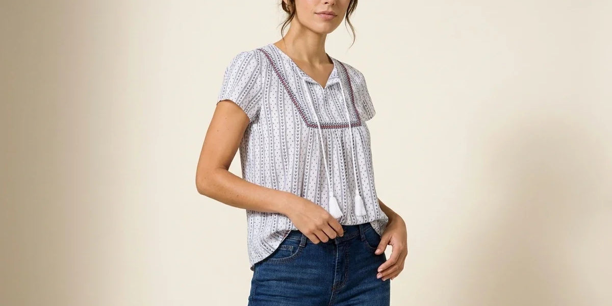 Brunette woman modeling a white short-sleeve peasant blouse with vertical blue dotted stripes and embroidered tassel ties, paired with dark blue jeans.