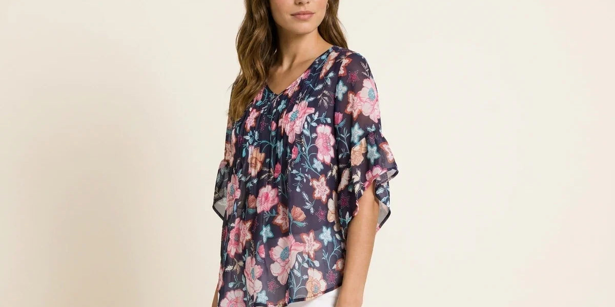 Brunette woman modeling a navy blue floral print chiffon blouse with tiered flutter sleeves paired with white pants.