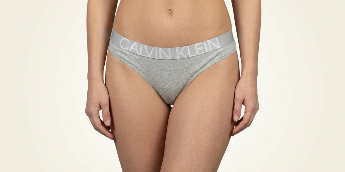 Cropped view of a woman wearing heather grey Calvin Klein cotton thong underwear with a signature logo elastic waistband.