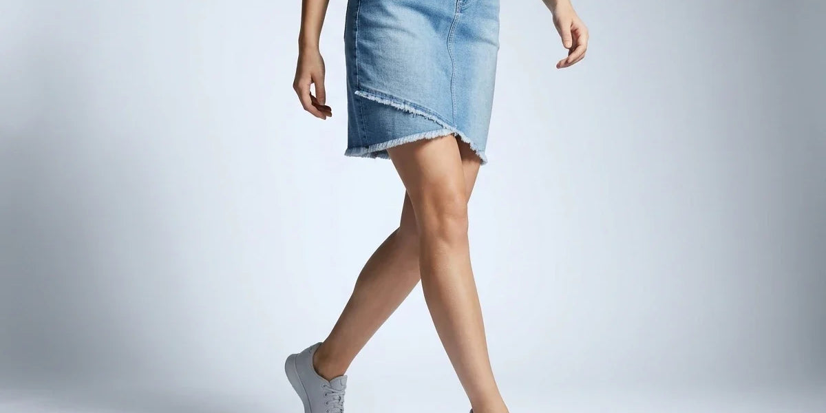 A model wearing a light blue denim asymmetrical mini skirt with frayed edges, paired with a white top and gray sneakers, captured against a light gray studio background.