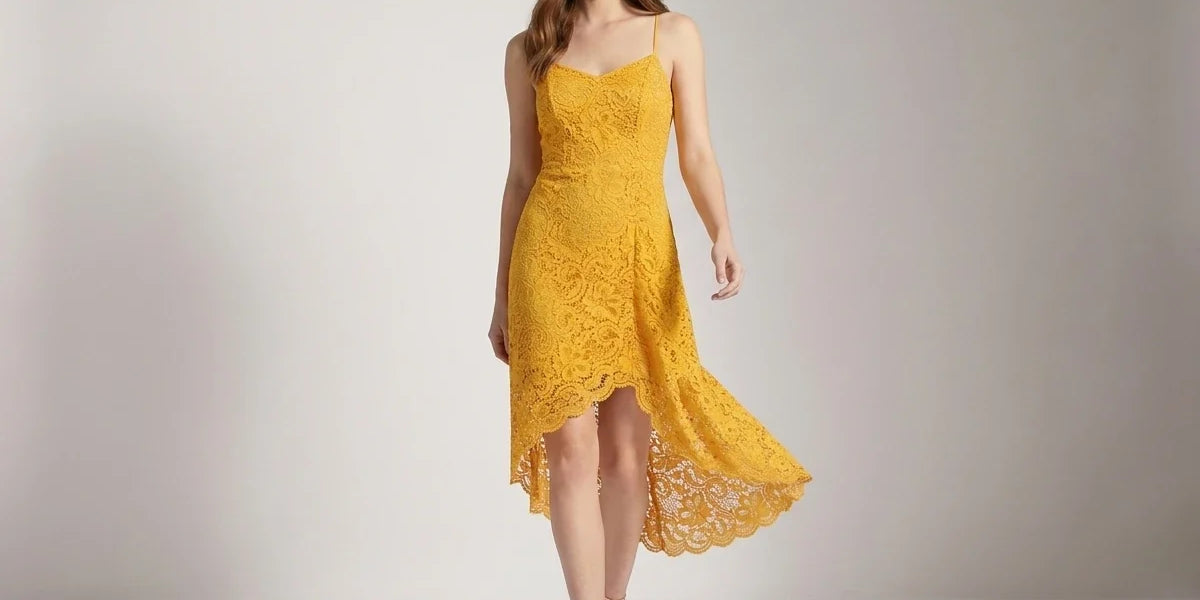 A woman modeling a bright yellow lace slip dress with spaghetti straps and a high-low hemline, standing against a neutral light gray studio backdrop.