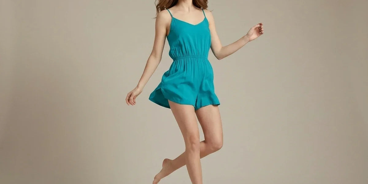 A woman wearing a bright turquoise sleeveless romper jumps barefoot against a plain beige studio background.