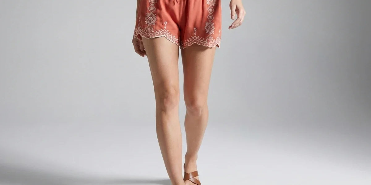 A model is shown from the waist down wearing coral pink embroidered shorts with a drawstring waist and scalloped lace trim, paired with brown slide sandals on a light gray studio background.