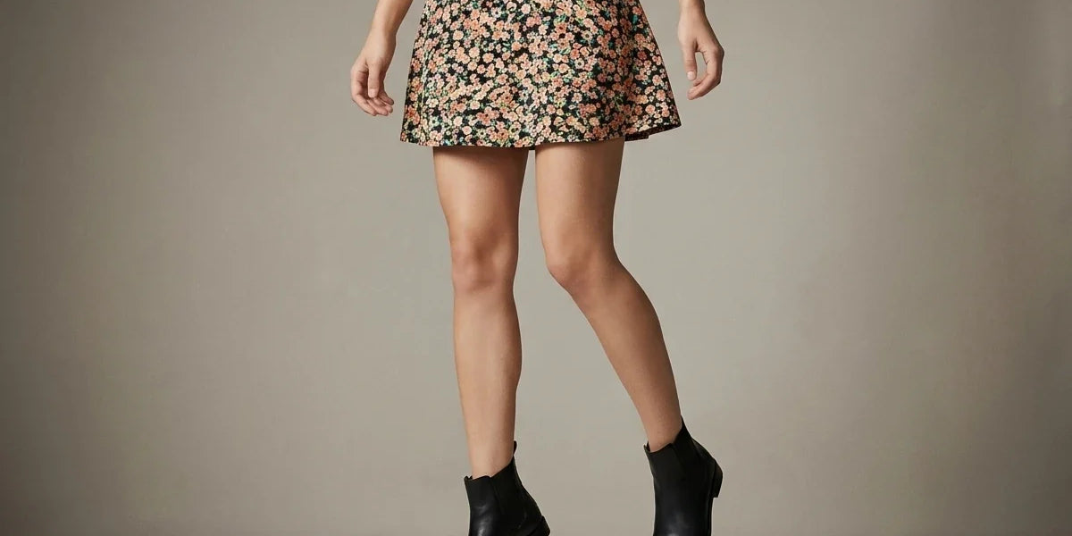 A model wearing a short A-line skirt with a dense floral print in pink, orange, and green on a dark background, paired with black ankle boots, captured mid-step against a neutral studio backdrop.