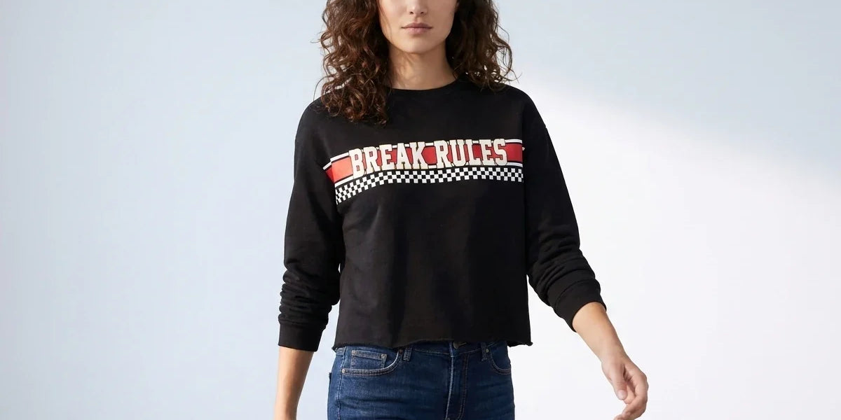 A woman modeling a black long-sleeve cropped sweatshirt with a red and white 'BREAK RULES' graphic across the chest, paired with blue jeans, against a light gray studio background.