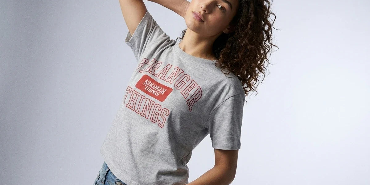 A woman modeling a heather gray graphic t-shirt with a red and white 'Stranger Things' print, paired with blue jeans, against a light gray studio background.