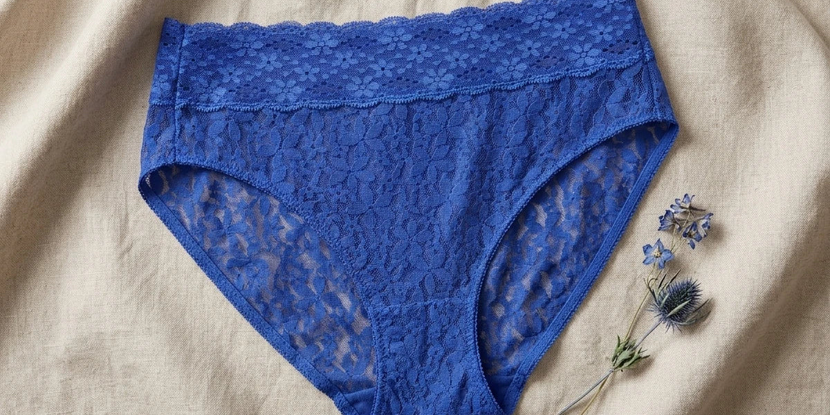 Blue lace high-waisted briefs laid flat on a beige linen fabric background, styled with small dried blue flowers in the corner.