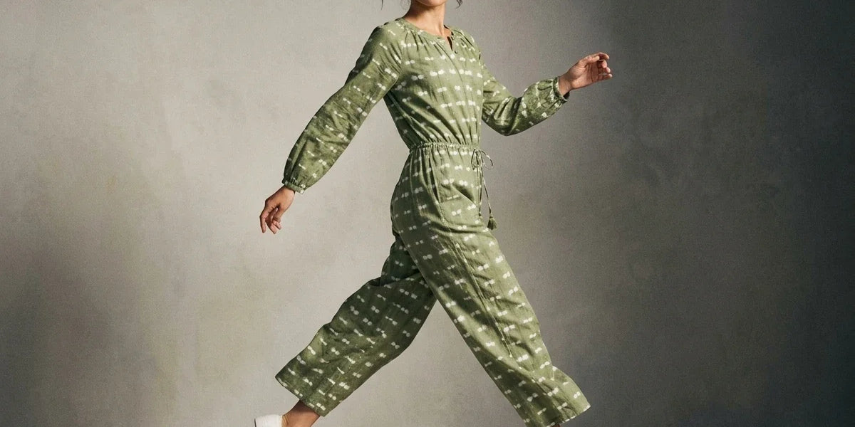 A woman wearing a sage green long-sleeve jumpsuit with a white geometric pattern and white sneakers, captured in a dynamic pose against a textured gray studio backdrop.