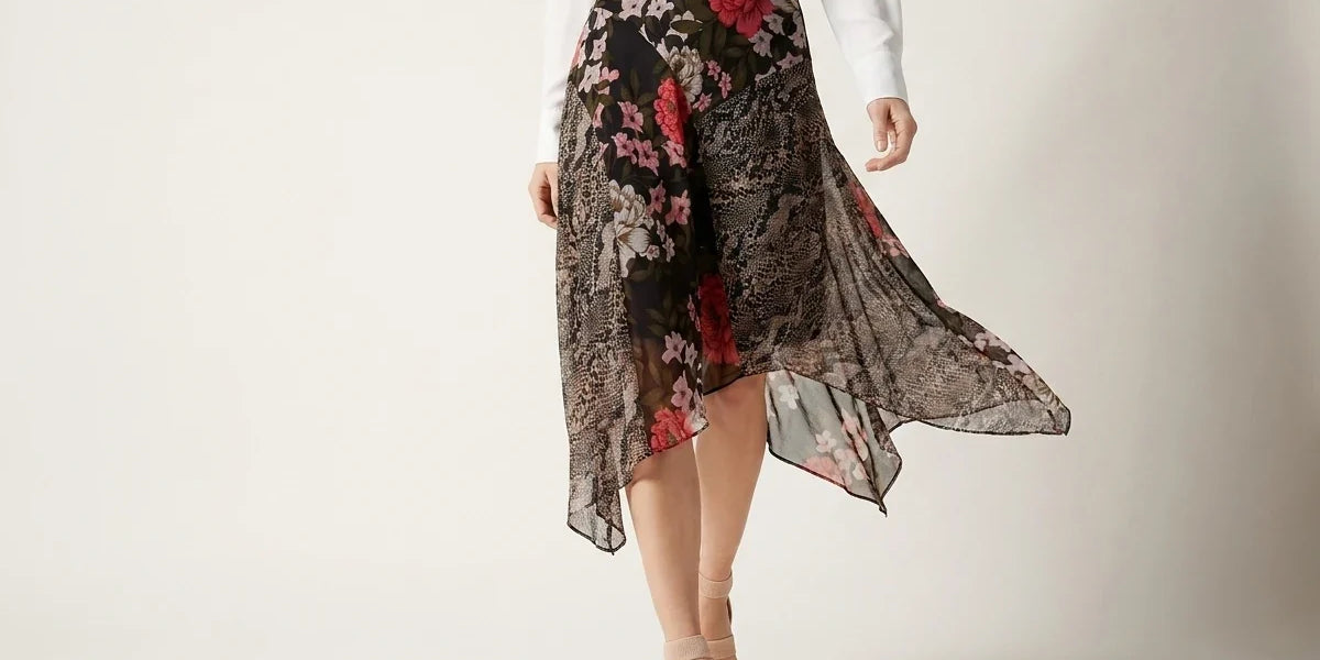 A model wearing a black high-waisted midi skirt with a red and pink floral lace overlay, paired with a white long-sleeve top and beige heeled sandals, set against a plain light gray studio background.