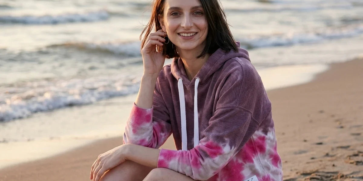 A woman sitting on a sandy beach at sunset wearing a pink and purple tie-dye hoodie with white drawstrings and a small Calvin Klein logo patch near the hem.
