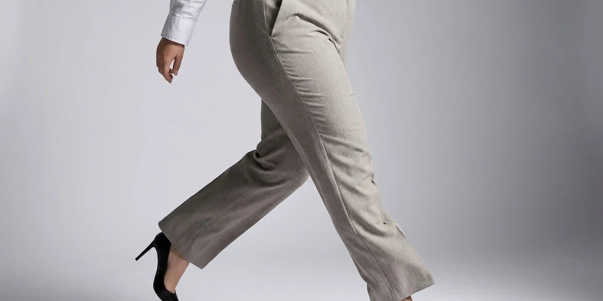 A person wearing light beige straight-leg trousers, a white long-sleeve blouse, and black pointed-toe heels, captured mid-step against a neutral gray studio background.