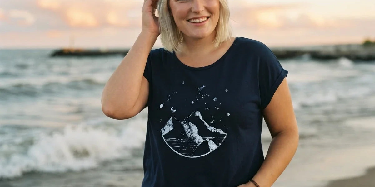 A smiling blonde woman wearing a navy blue graphic t-shirt with a white moon and mountain design, standing on a beach during sunset.