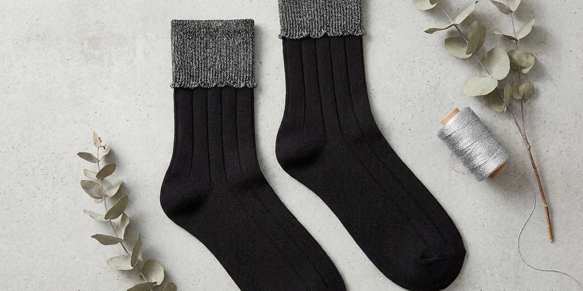 "Flat lay of women's black ribbed crew socks with metallic silver folded cuffs, styled with eucalyptus branches and silver thread spool.