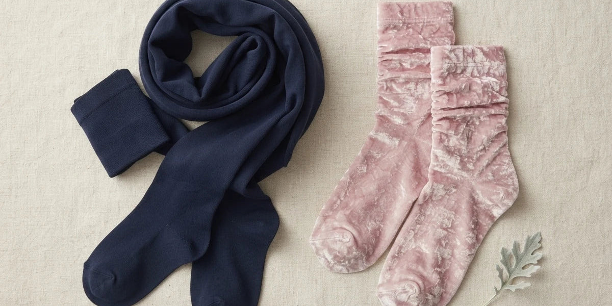 A navy blue scarf and a pair of dusty pink velvet socks arranged on a textured beige fabric background with a small decorative leaf.