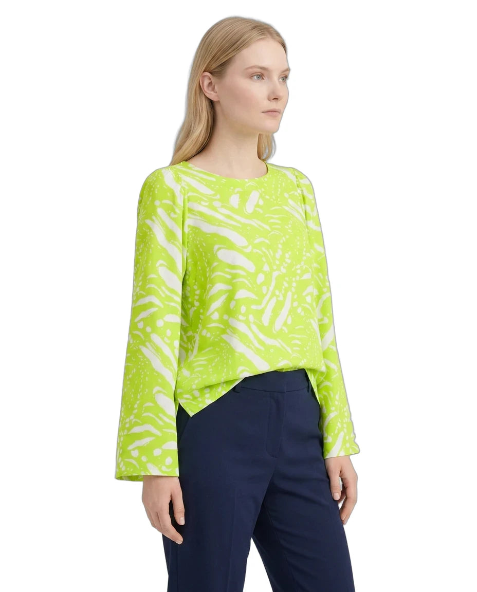 A woman modeling a bright lime green long-sleeve blouse with an abstract white brushstroke pattern, paired with navy blue trousers, against a plain white background.