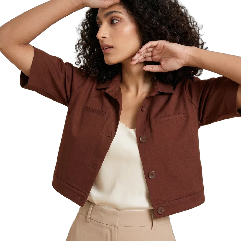 A woman wearing a cropped brown short-sleeve button-front jacket over a cream top and beige trousers, posed against a plain white studio background.