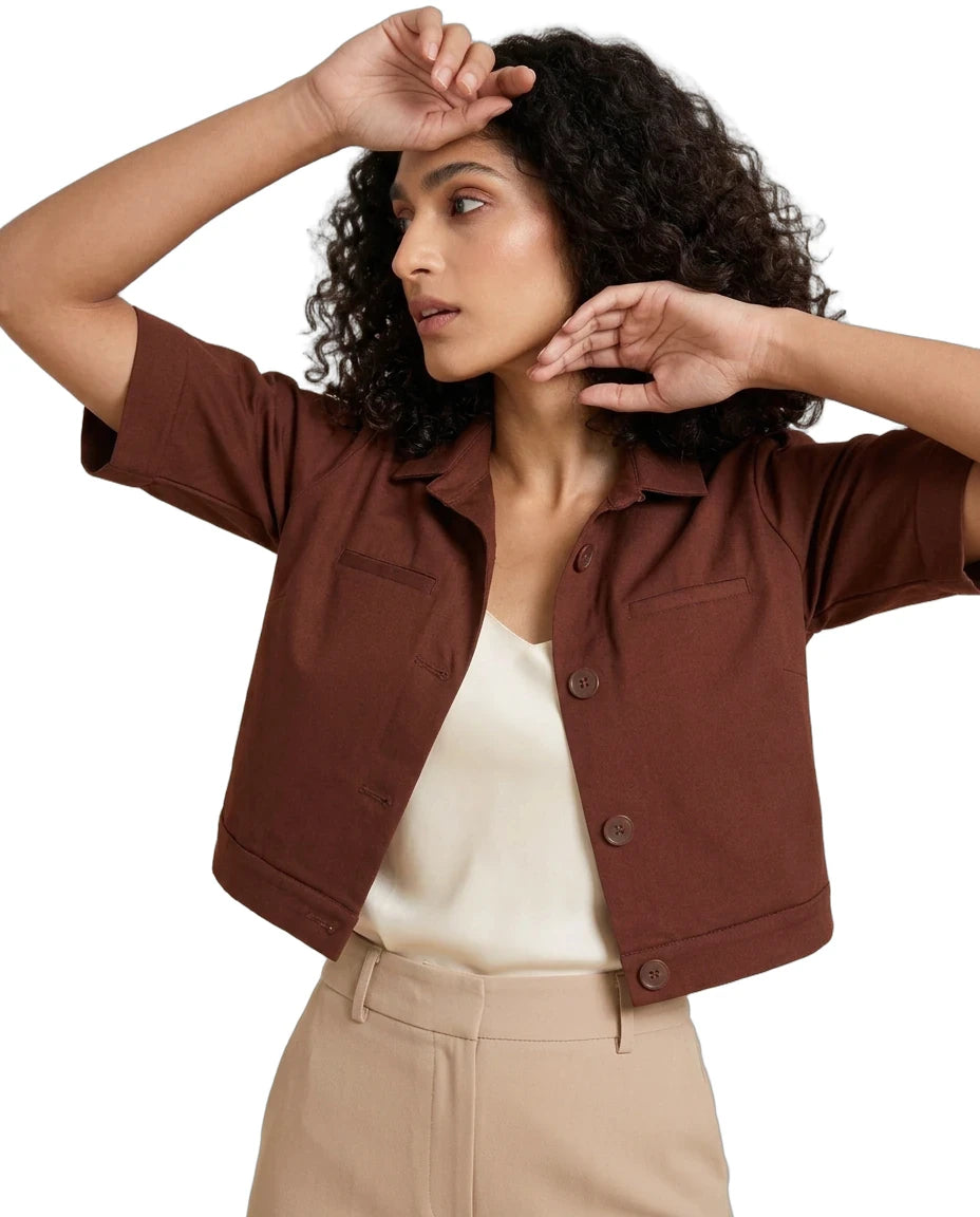 A woman wearing a cropped brown short-sleeve button-front jacket over a cream top and beige trousers, posed against a plain white studio background.