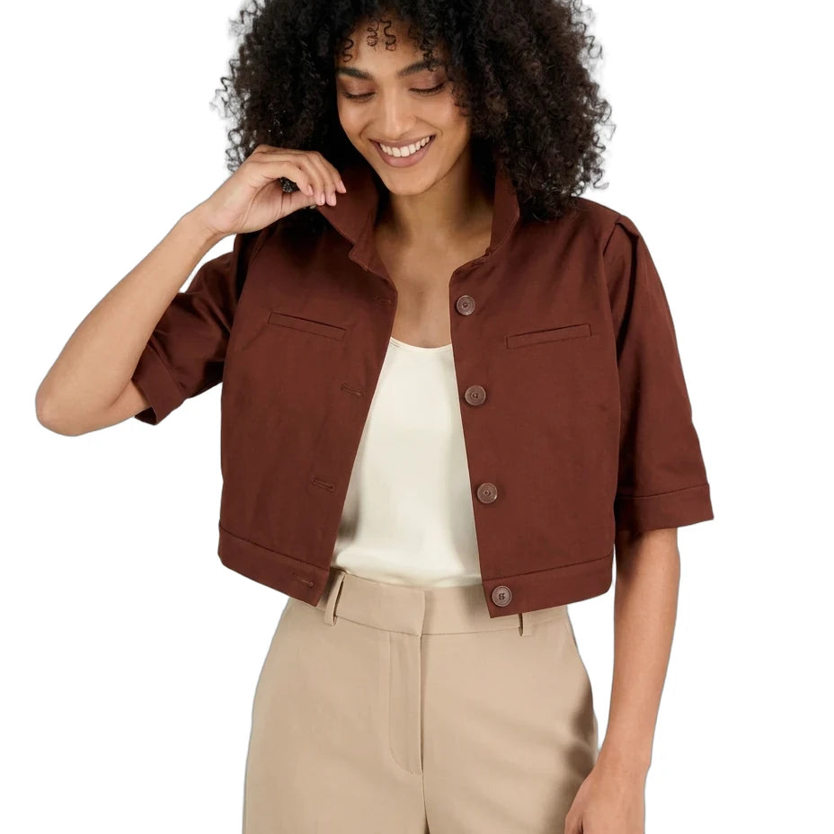 A woman with curly black hair wearing a brown cropped short-sleeve button-up jacket over a cream top and beige trousers, photographed against a plain white background.