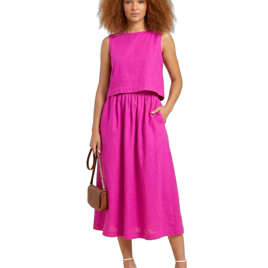 A woman modeling a bright pink two-piece linen set consisting of a sleeveless cropped top and a matching midi skirt with pockets, styled with a brown handbag and white strappy heels against a white background.
