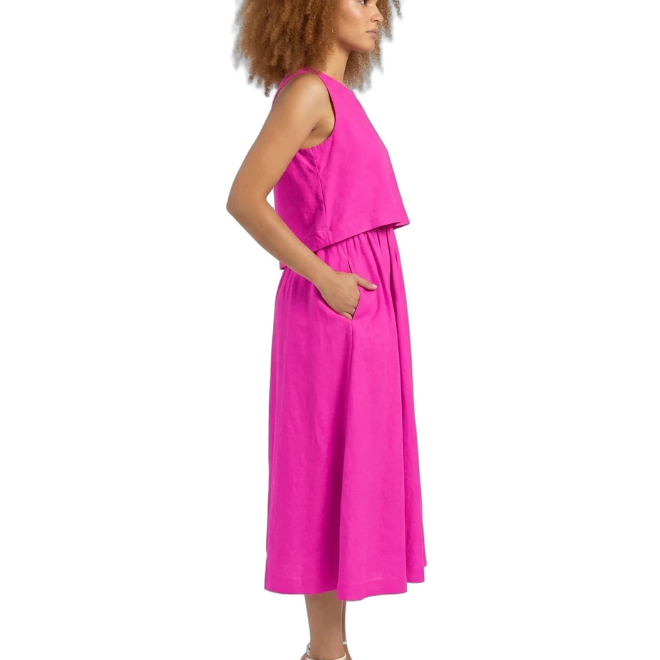 A woman shown in side profile wearing a sleeveless bright pink midi dress with a draped yoke and side pockets, paired with white strappy high-heeled sandals, against a plain white background.