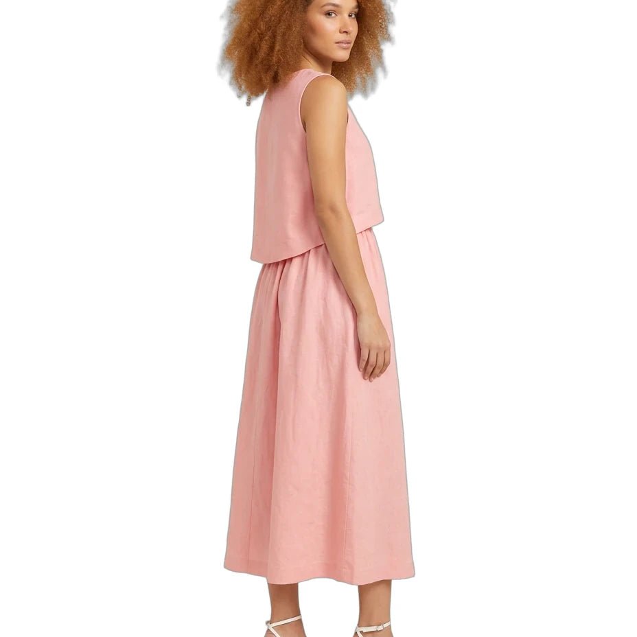 A woman with curly light brown hair is shown from the side wearing a sleeveless dusty pink midi dress with a layered ruffle top and white strappy high-heeled sandals, set against a plain white background.