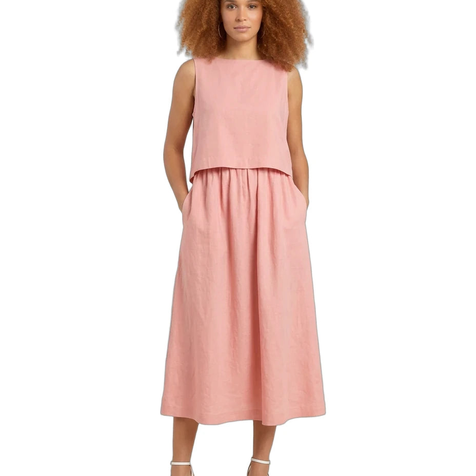 A woman with voluminous curly brown hair wearing a sleeveless dusty pink two-piece linen set consisting of a cropped top and a matching midi skirt with pockets, paired with white strappy high-heeled sandals, set against a plain white background.