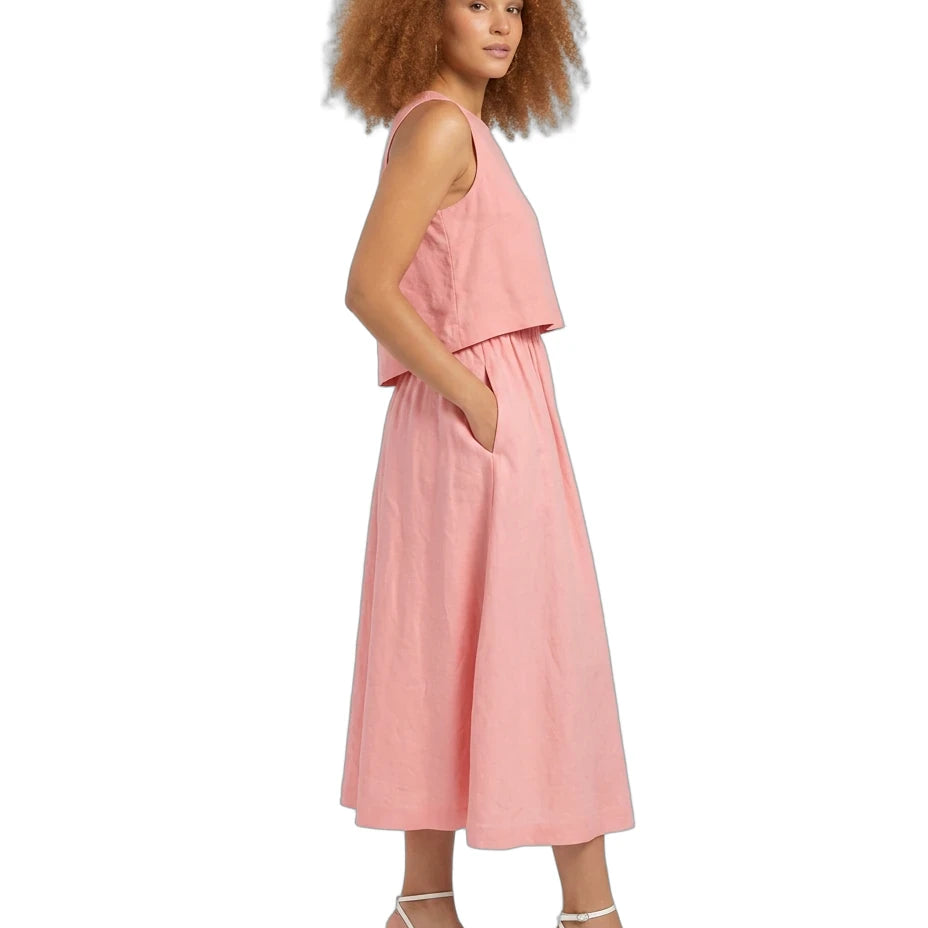 A woman with curly light brown hair is shown in profile wearing a sleeveless dusty pink linen midi dress with a ruffled overlay and side pockets, paired with white strappy block-heel sandals, against a solid white background.
