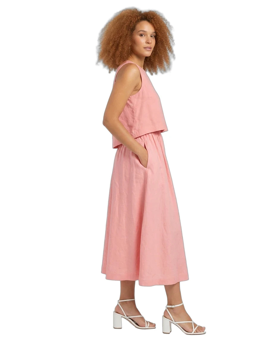 A woman with curly light brown hair is shown in profile wearing a sleeveless dusty pink linen midi dress with a ruffled overlay and side pockets, paired with white strappy block-heel sandals, against a solid white background.