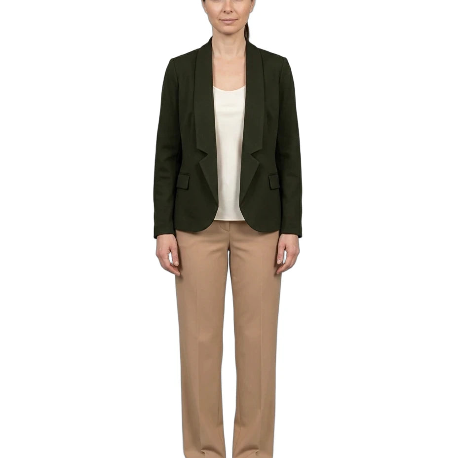 A woman stands against a plain white background wearing a dark olive green blazer over a cream top, beige trousers, and white sneakers.