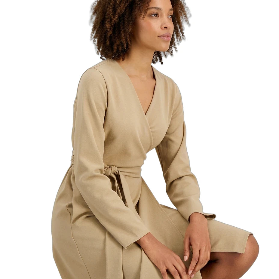 A woman with curly brown hair wearing a beige wrap dress with long sleeves and a tie waist, posing against a solid white background.