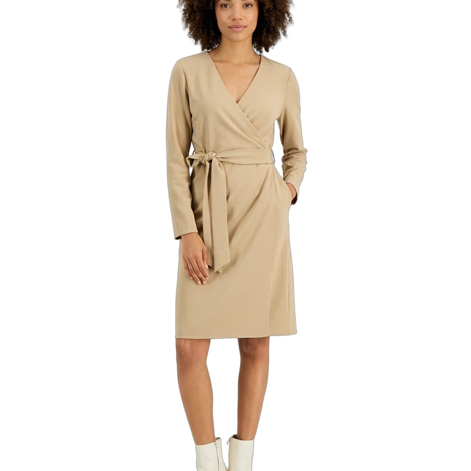 A woman modeling a beige wrap dress with long sleeves and a matching waist tie, paired with white ankle boots, against a plain white background.