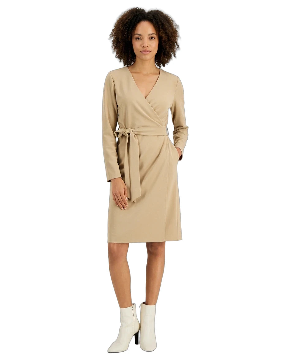 A woman modeling a beige wrap dress with long sleeves and a matching waist tie, paired with white ankle boots, against a plain white background.