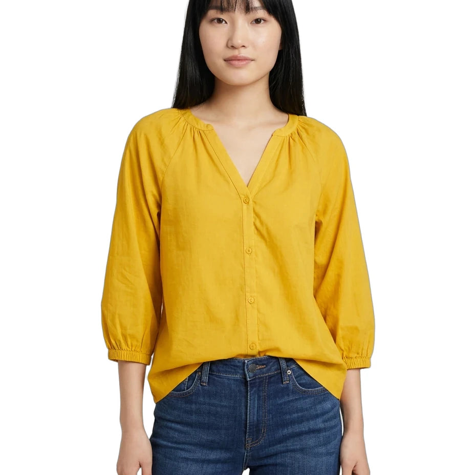 A woman wearing a mustard yellow blouse with three-quarter puff sleeves and a gathered V-neckline, paired with dark blue jeans, against a plain white background.