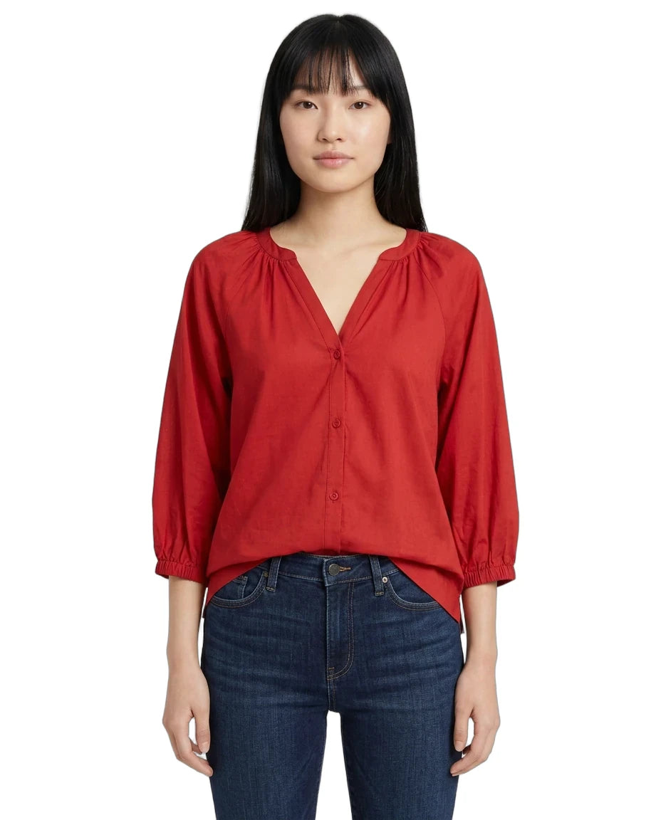 A woman modeling a red long-sleeve blouse with a gathered V-neckline and button front, paired with dark blue jeans, against a plain white background.