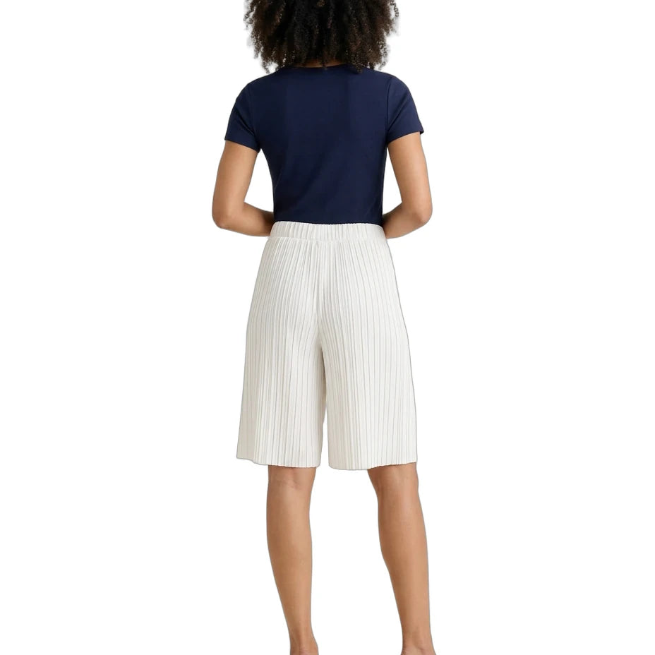 A woman shown from the back wearing a navy blue short-sleeve top, off-white pleated knee-length shorts, and tan block-heel mules against a plain white background.