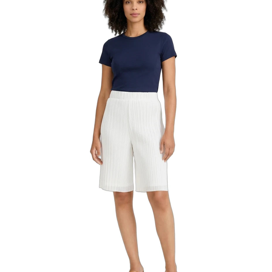 A woman modeling a navy blue short-sleeve crewneck top and white pleated knee-length shorts, paired with tan heeled mules, against a plain white background.