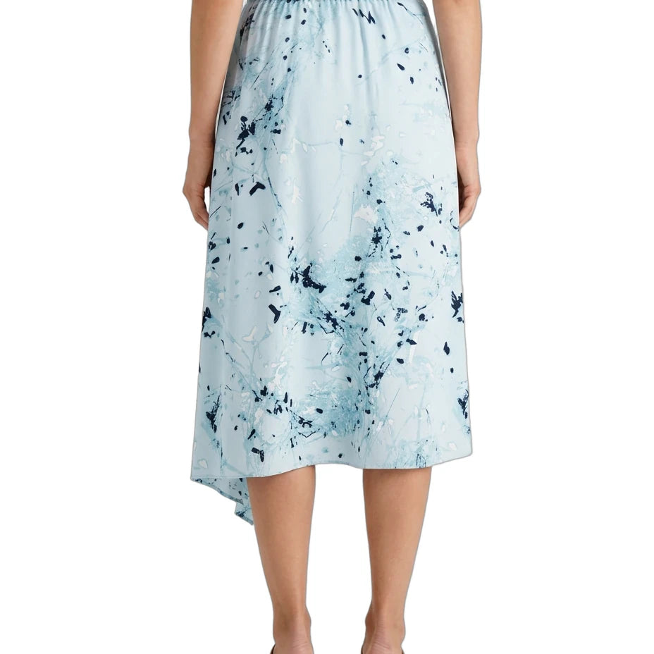 A model is shown from the back wearing a light blue midi skirt with a dark blue abstract paint splatter pattern, paired with a navy top and tan block-heel sandals, against a plain white background.
