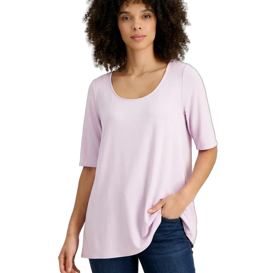 A woman with curly dark hair wearing a light lavender scoop-neck t-shirt and dark blue jeans, standing against a plain white background.