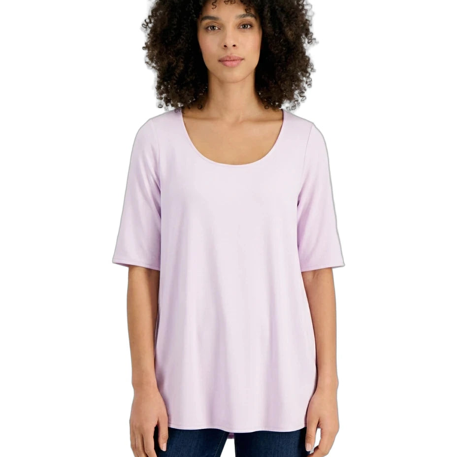 A woman with curly dark hair wearing a light lavender short-sleeve scoop-neck top and dark blue jeans, standing against a plain white background.