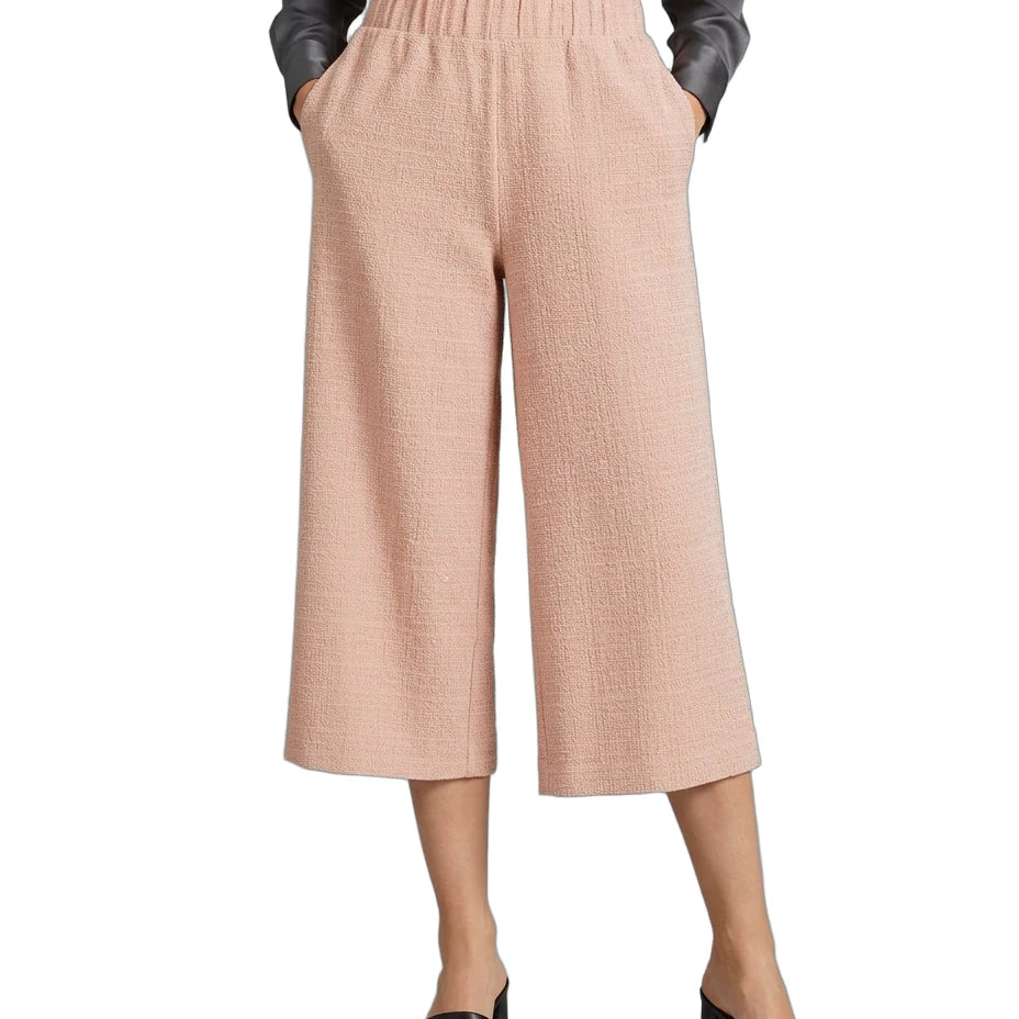 A model wearing a pair of light peach-pink cropped wide-leg pants with a textured weave, paired with a dark gray button-up shirt and black slide sandals, shown against a plain white background.