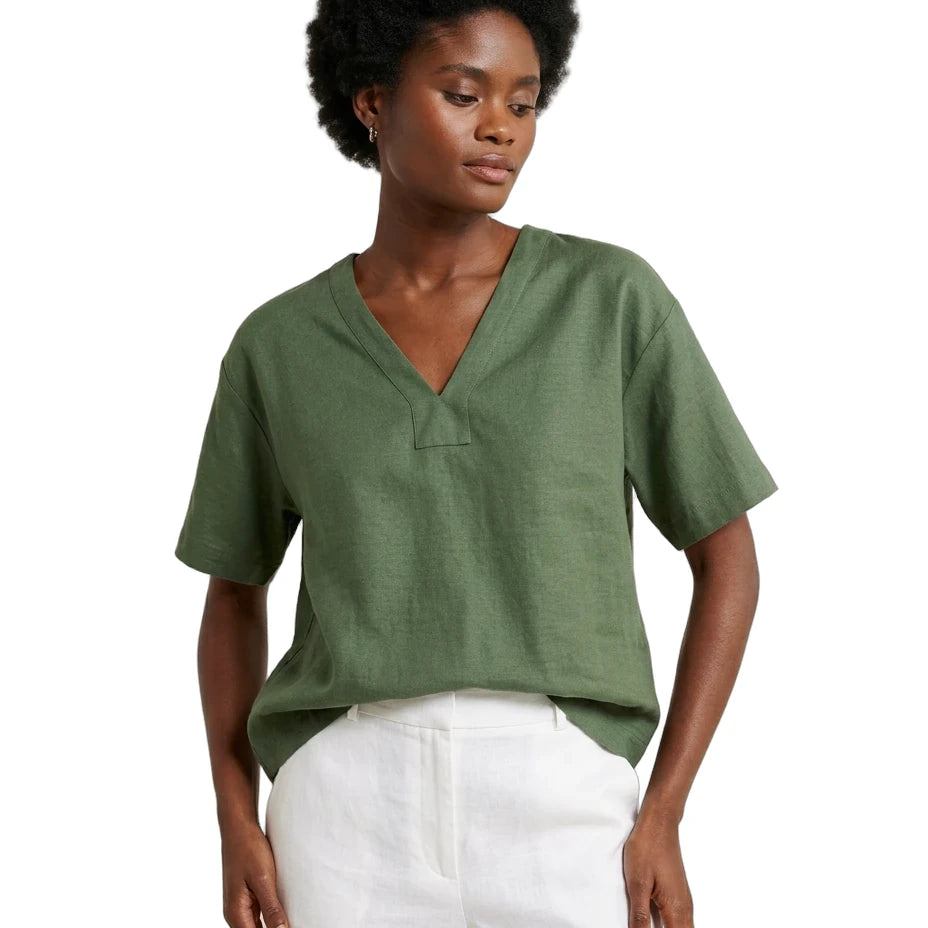 A woman wearing a short-sleeve olive green V-neck top and white pants, photographed against a plain white studio background.