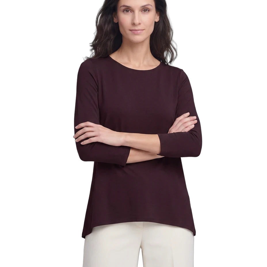 A woman modeling a dark plum long-sleeve top with a crew neckline, paired with off-white pants, against a plain white background.
