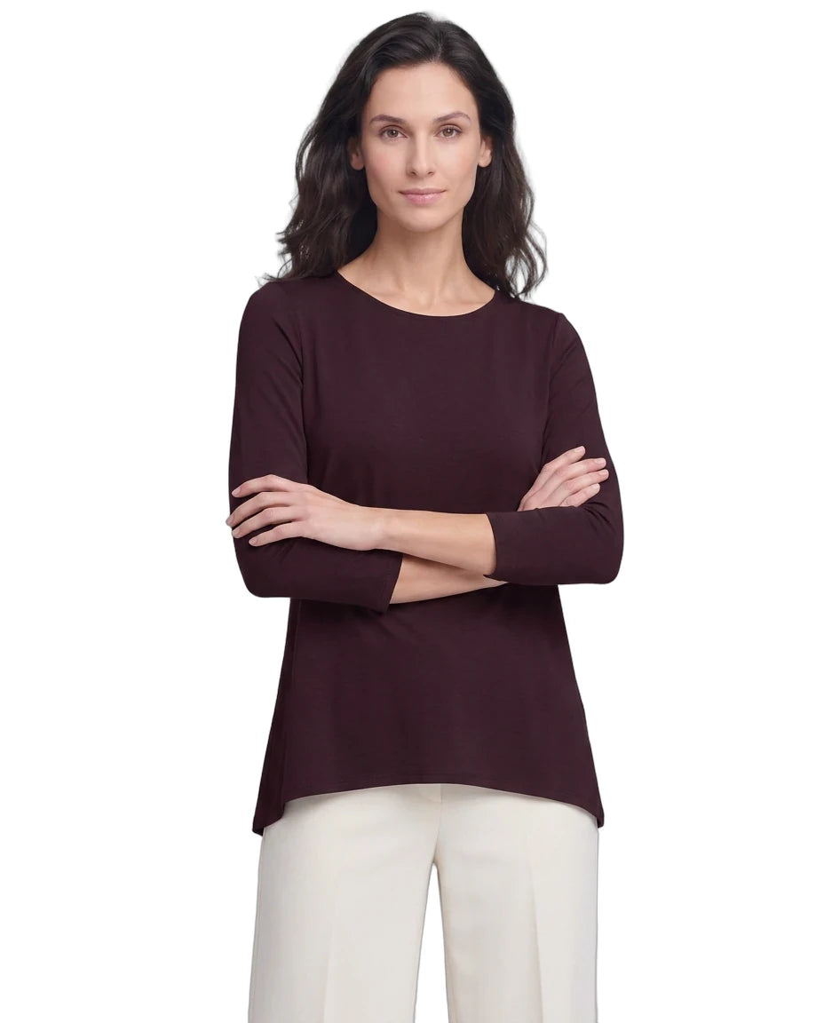 A woman modeling a dark plum long-sleeve top with a crew neckline, paired with off-white pants, against a plain white background.
